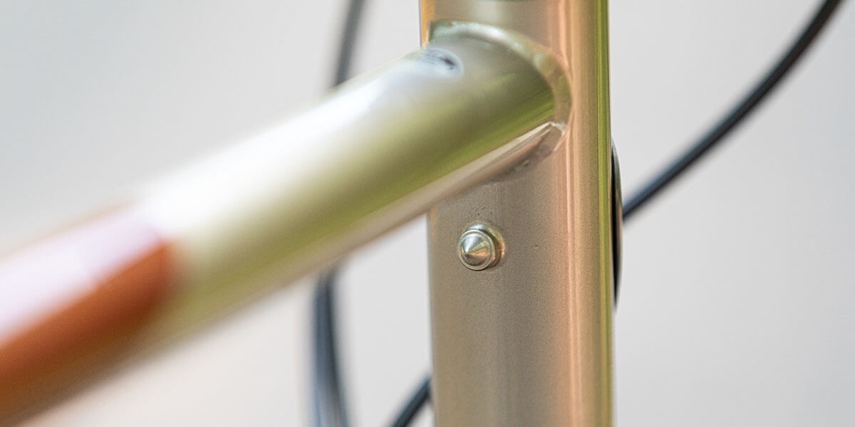 All-City gold Spacehorse bike, close up of top tube and heat tube junction with pump peg with an outdoor background