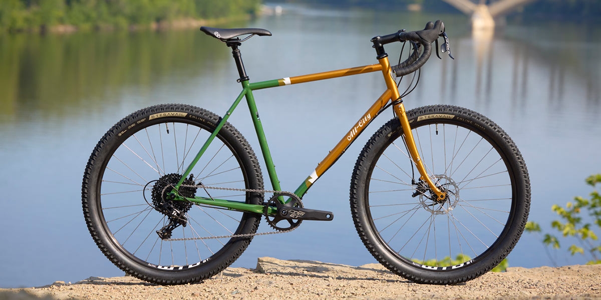 All-City Gorilla Monsoon GRX complete bike in Tangerine Evergreen color, side view with river in background