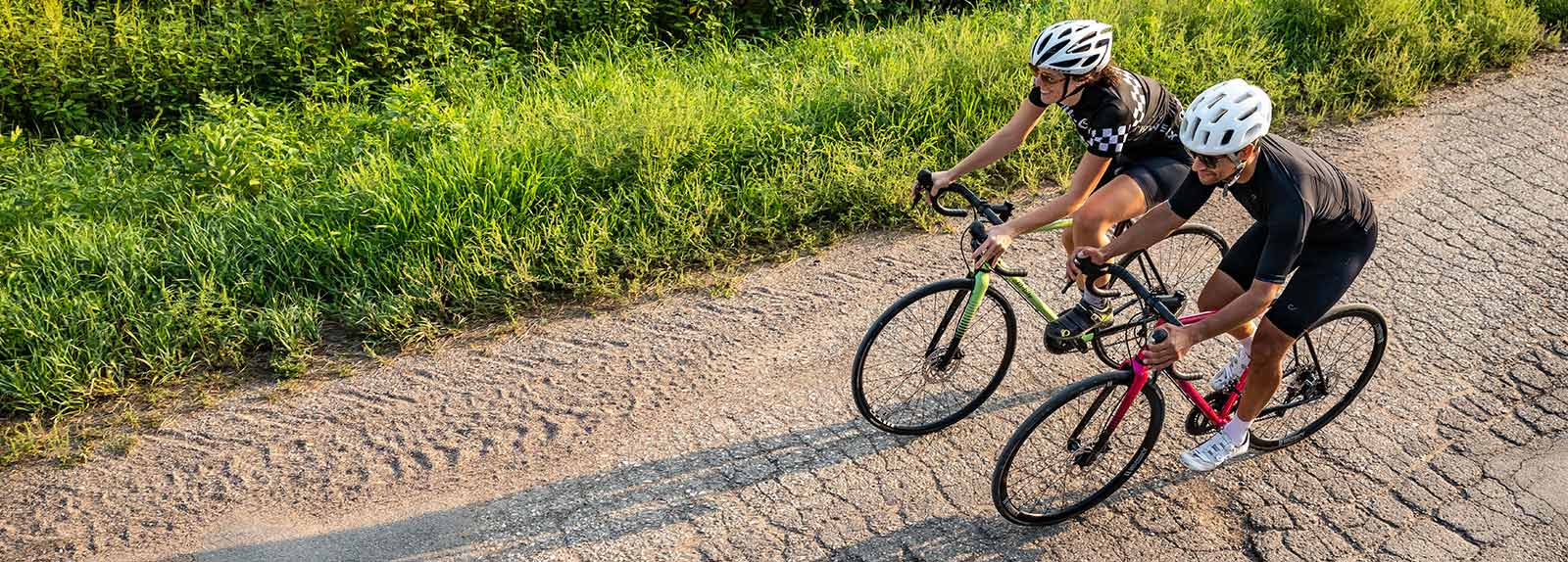 Two cyclists on rough asphalt road with tall grass on side in cycling helmets and apparel riding All-City Zig Zag bikes