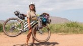 Kae-Lin on gravel road smiling on sunny day, holding loaded Gorilla Monsoon bike off ground, mountains in background