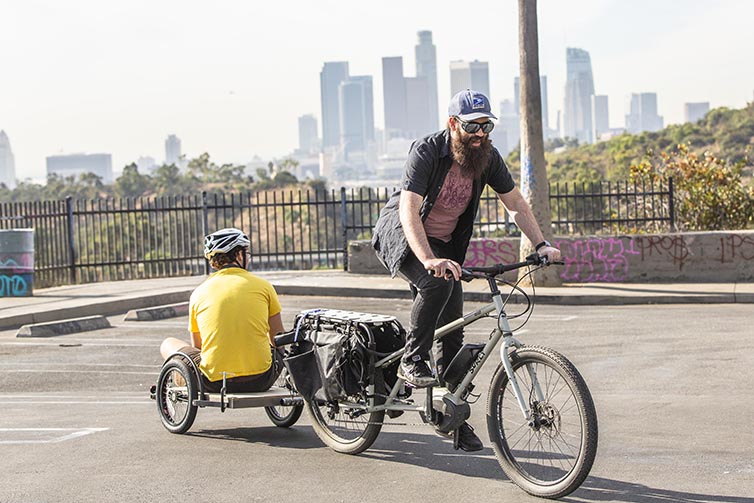 Person on bike pulls another person sitting on attachment in front of LA skyline