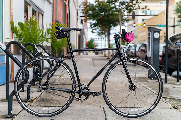 Dark gray/black All-City Big Block on bike rack on sidewalk with small pink handlebar bag