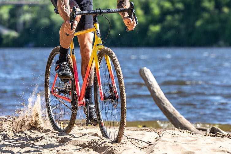 Person riding Nature Cross Single Speed Pink Lemonade bike on sandy river shoreline