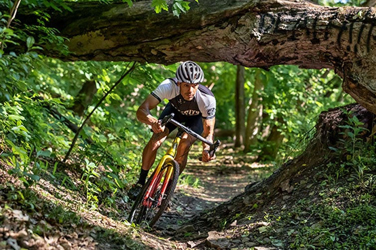 Person in helmet and cycling apparel riding Nature Cross Single Speed bike on forest trail ducking fallen tree