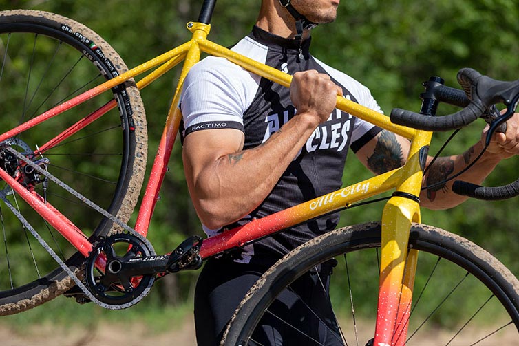 Person in All-City jersey shouldering Nature Cross Single Speed bike, showing bike side view, sand on tires