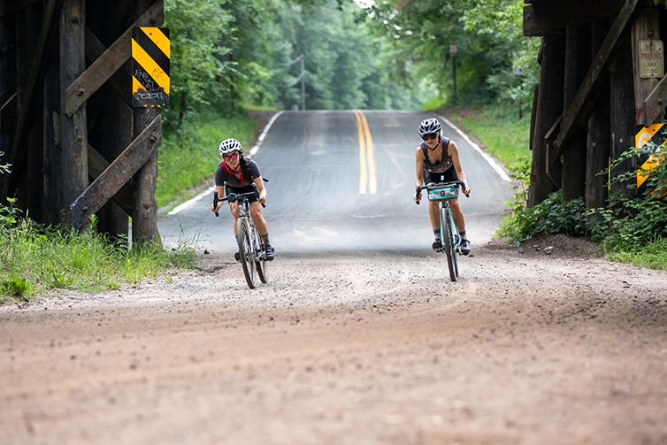 Two cyclists riding Space Horse bikes wearing sunglasses, transitioning from paved road to gravel road under bridge