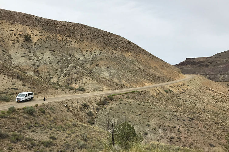 White Sprinter van parked on side of gravel desert road with photographer crouching, looking through camera on tripod