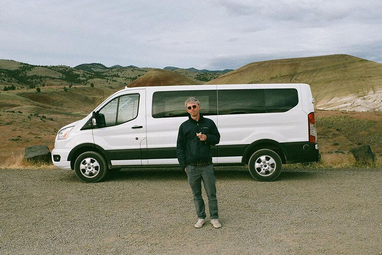 Mike wearing sunglasses, dark jacket, and jeans standing in front on Sprinter van eating snack on desert road