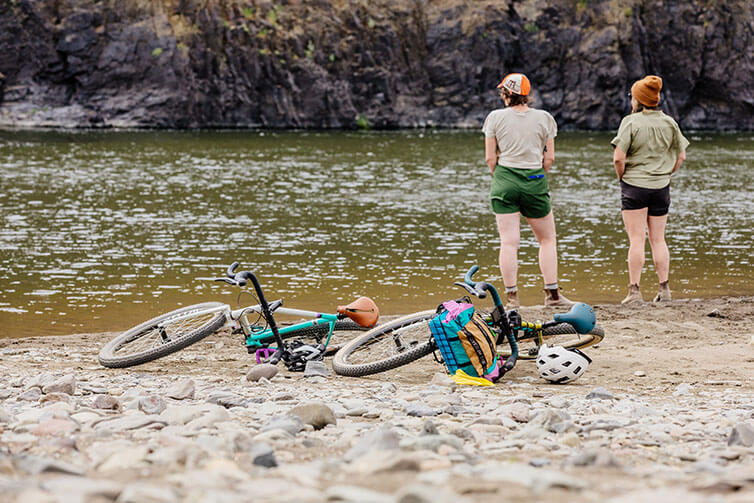 Alix and Wang standing on river shore, Gorilla Monsoon bikes laying with gear and helmets