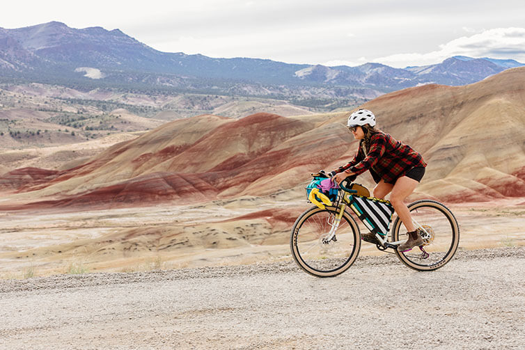 Wang out of saddle riding loaded Gorilla Monsoon, descending gravel road with painted hills and mountains in background