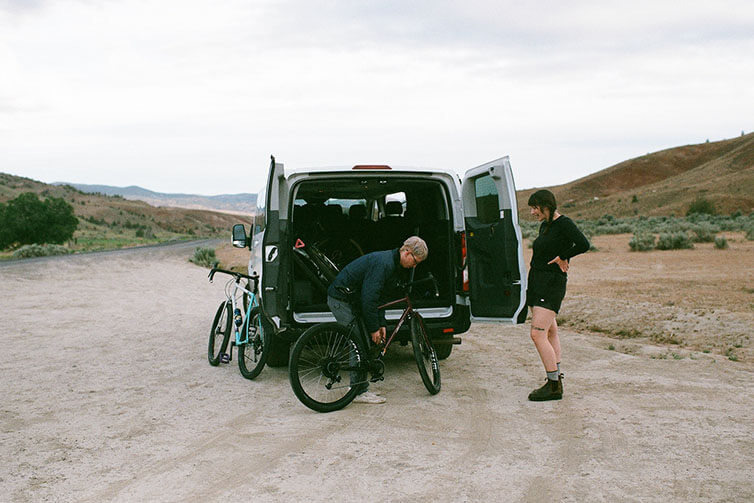 Sprinter van parked on desert road, back doors open and Mike adjusting Gorilla Monsoon bike while talking with Alix