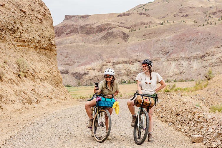 Wang and Alix stopped on mountain gravel road with leg over loaded Gorilla Monsoon bikes, taking a selfie break