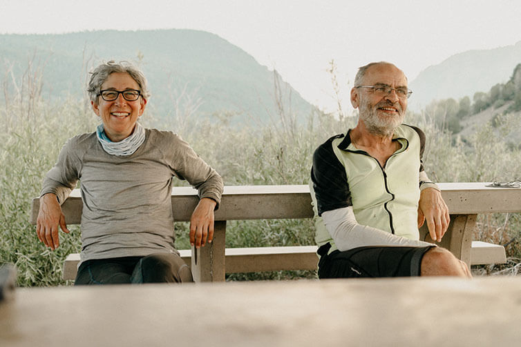 Jeri and Charles smiling sitting on bench, tall grass an mountains in background