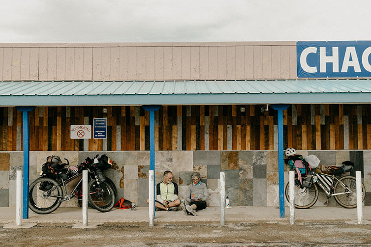 Jeri and Charles sitting under awning of building out of rain looking at map, loaded bikes parked