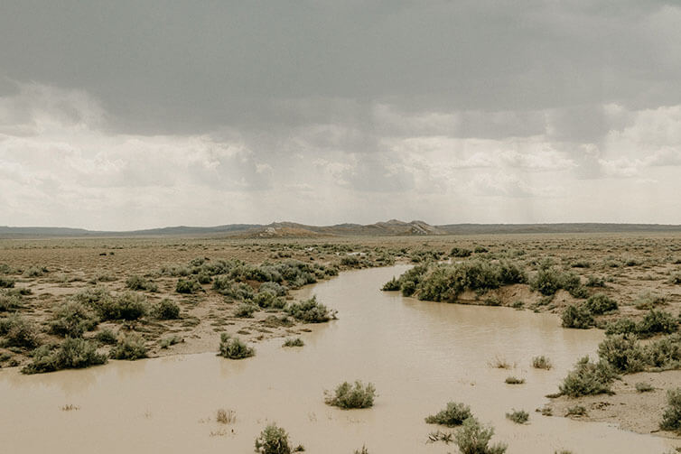 Muddy river lined with sage brush weaving through high plains, rain clouds overhead