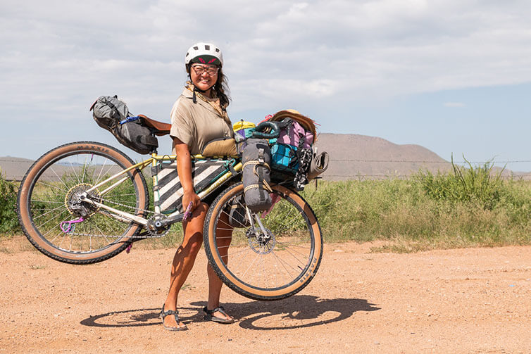 Kae-Lin on gravel road smiling on sunny day, holding loaded Gorilla Monsoon bike off ground, mountains in background