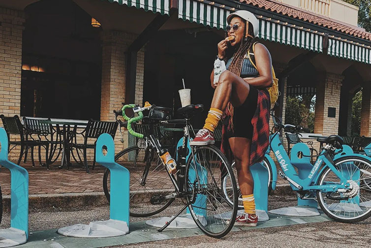 Person standing next to parked bike with foot resting atop rear wheel while eating snacks