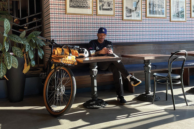 Person sitting inside coffee shop looking at phone, bike parked at talbe with Beatbox Front Rack Bag on bike