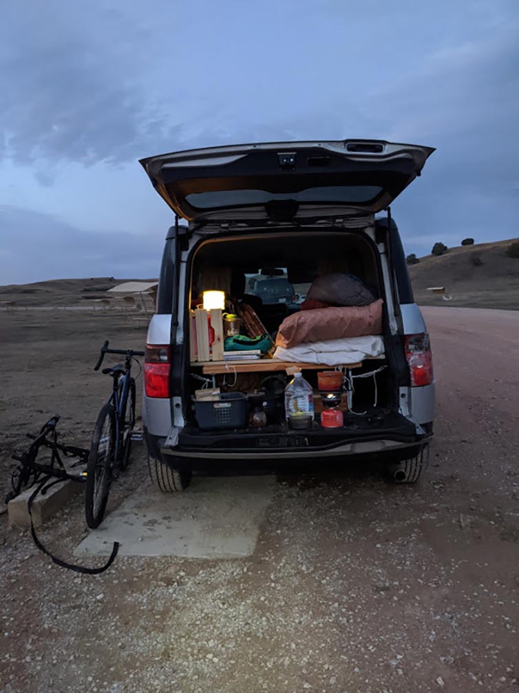 Van with back hatch open with sleeping and camping gear inside and bike leaning against at sunset