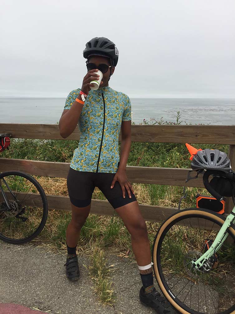 Grace wearing helmet and cycling apparel drinking out of paper cup in front of fence with bikes parked, ocean in background