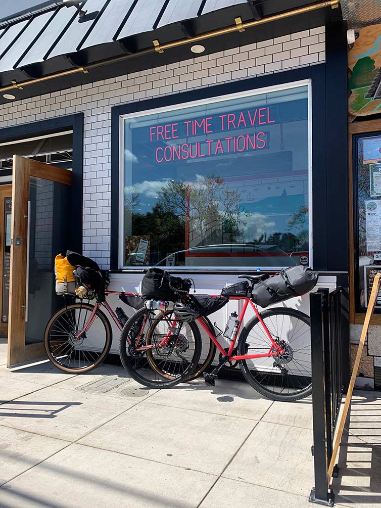 Two loaded bikes leaning up against white building with large window neon sign “Free Time Travel Consultations”
