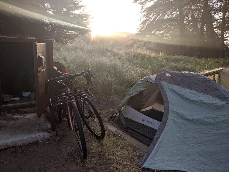 Sun rising on camp, two tents with bikes parked against iron stove