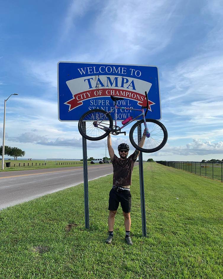 Cyclist wearing helmet and sunglasses standing, holding bike overhead, in front of Tampa city sign next to road