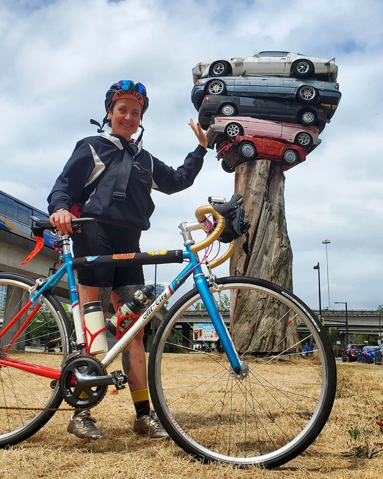 Person standing outside holding bike with one hand, other hand looking to hold up or push five cars stacked on dead tree