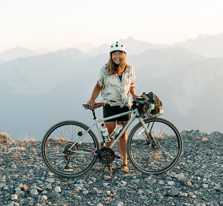 Karen Wang with her bike atop a mountain pass, mountain tops in background