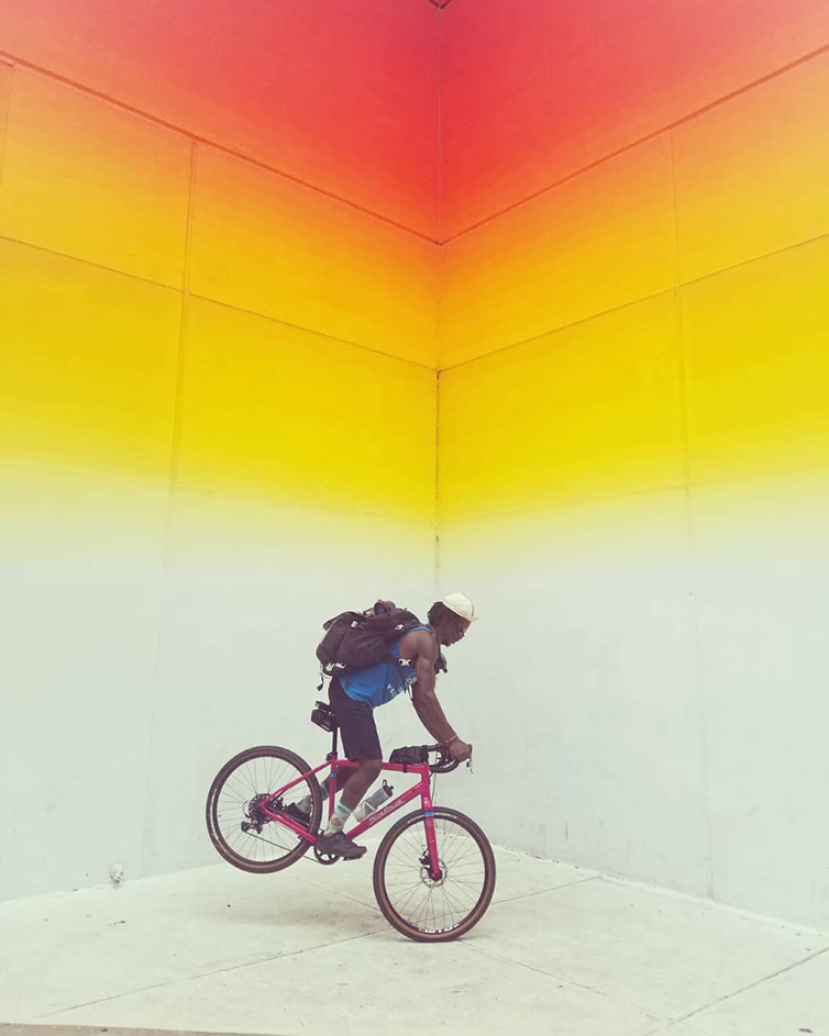 Person on pink bike, holding front brake to balance on front wheel, concrete walls painted, orange to white fade