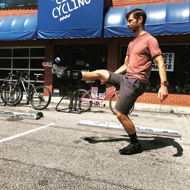 Person in front of bike shop with blue awning, bike racks out front, kicking hacky sack by themselves in parking lot