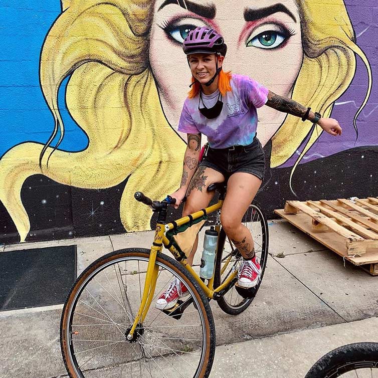 Person wearing bicycle helmet on bike, balancing, doing track stand, with one hand on handlebar in front of mural