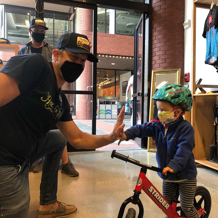 Person wearing mask, trucker hat, kneeling in bike shop giving high-five to child on balance bike wearing helmet and mask