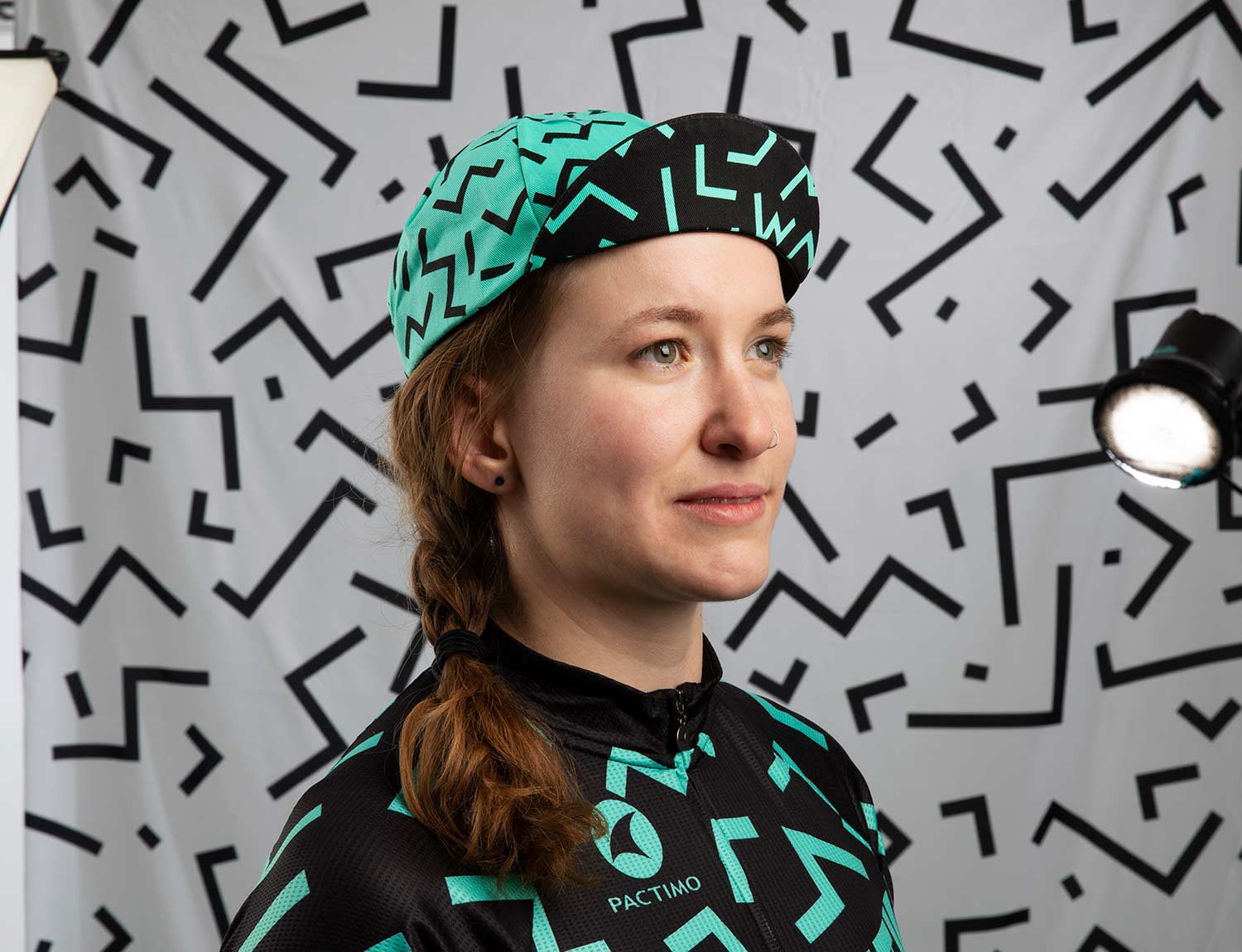 Person wearing All-City Cycles teal and black Max Cycling Cap in front of a grey abstract background