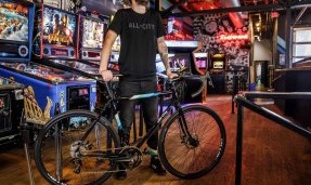 Person wears black All-City Cycles Travel Wool Shirt holding bike in front of arcade 