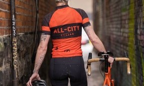 Back of a person wearing black and orange All-City classic jersey 2.0 walking down alley
