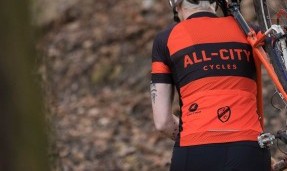 Back of a person wearing black and orange All-City classic jersey 2.0 walking through trail