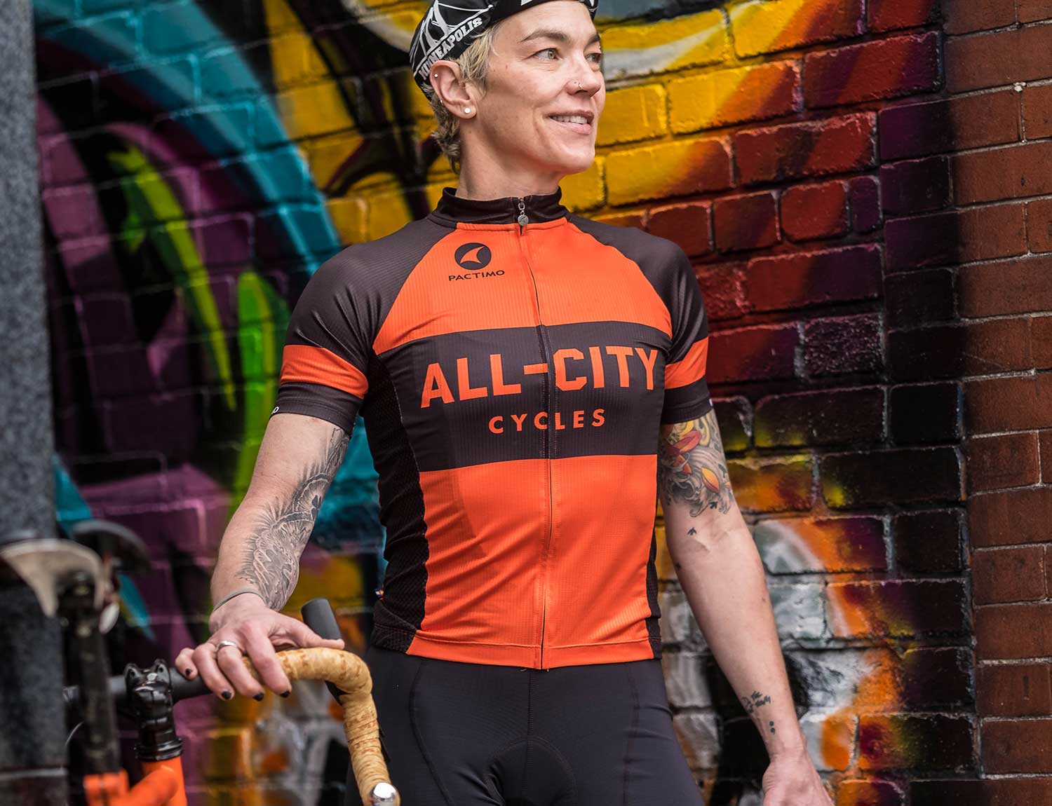 Person wearing black and orange All-City classic jersey 2.0 in front of graffiti wall
