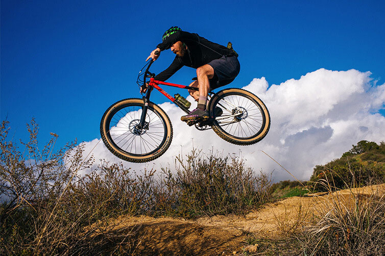 Mountain biker on All-City steel mountain bike catching air off jump blue sky and clouds in background