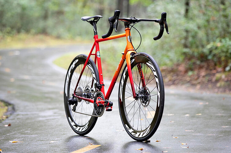 All-City Zig Zag steel road bike, orange, propped up on wet paved trail in forest