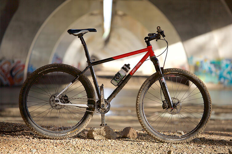 All-City Log Lady red and black steel mountain bike under bridge with river in background